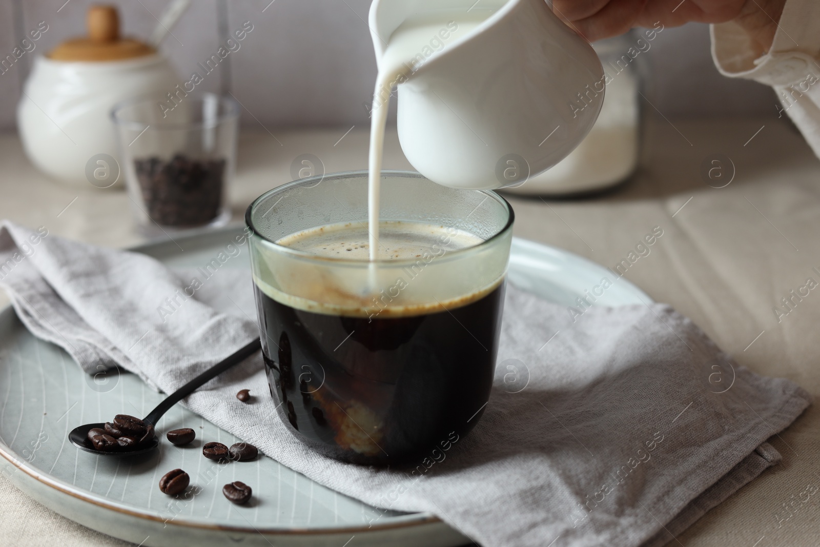 Photo of Woman pouring milk into cup with coffee at light table, closeup