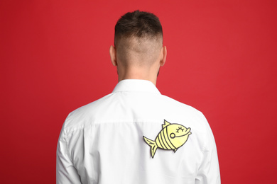 Photo of Man with paper fish on back against red background. April fool's day