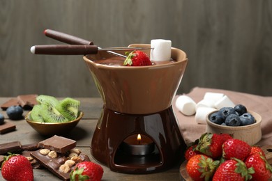 Photo of Fondue pot with melted chocolate, different fresh berries, kiwi, marshmallows and forks on wooden table