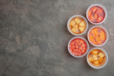 Plastic containers of tasty jelly desserts with fruit pieces on grey background, flat lay. Space for text