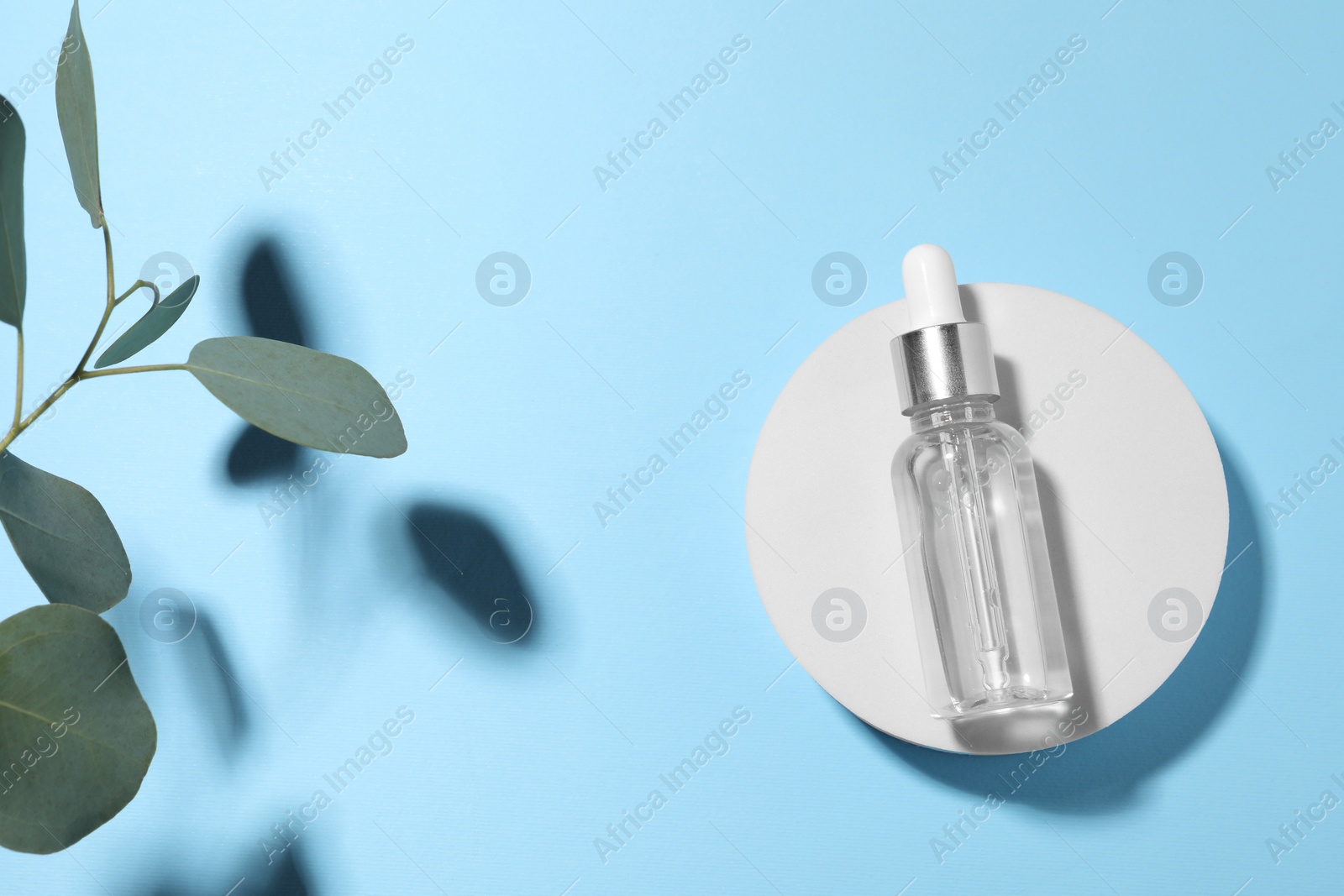 Photo of Bottle of cosmetic oil and green leaves on light blue background, top view