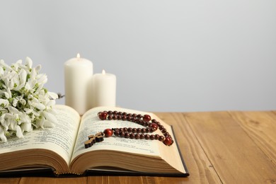 Bible, rosary beads, flowers and church candles on wooden table against light background. Space for text