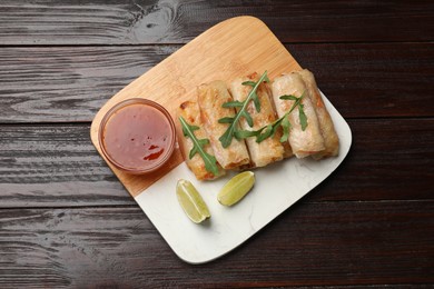 Photo of Tasty fried spring rolls, lime, arugula and sauce on wooden table, top view