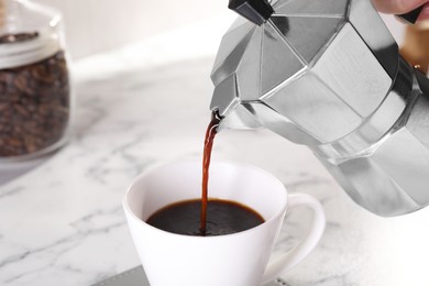 Photo of Pouring aromatic coffee from moka pot into cup at white marble table, closeup