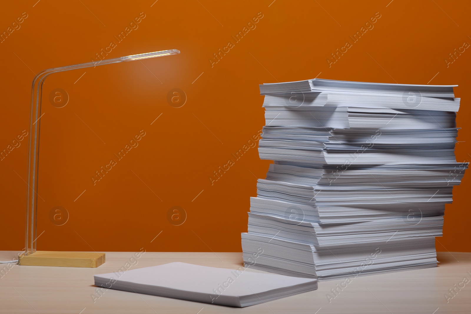 Photo of Stack of paper sheets and lamp on white wooden table near orange wall