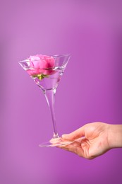 Photo of Woman holding martini glass with water and pink flower on violet background, closeup