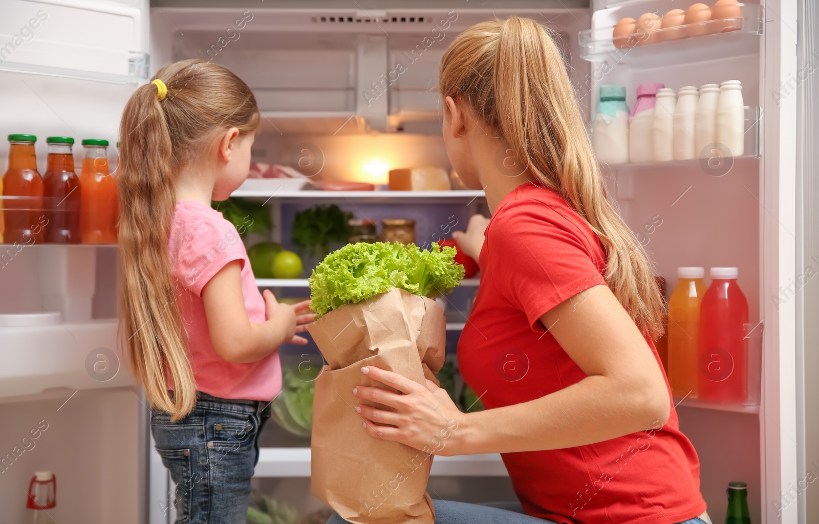 Photo of Young mother and daughter with paper bag full of food near refrigerator at home