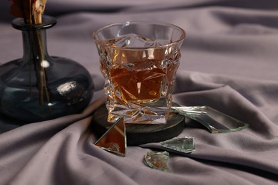Photo of Glass of tasty alcohol drink and shards of broken mirror on grey fabric