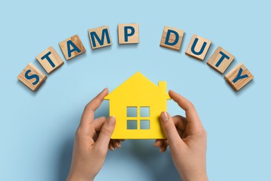 Image of Woman holding house figure and wooden cubes with text Stamp Duty on light blue background, top view