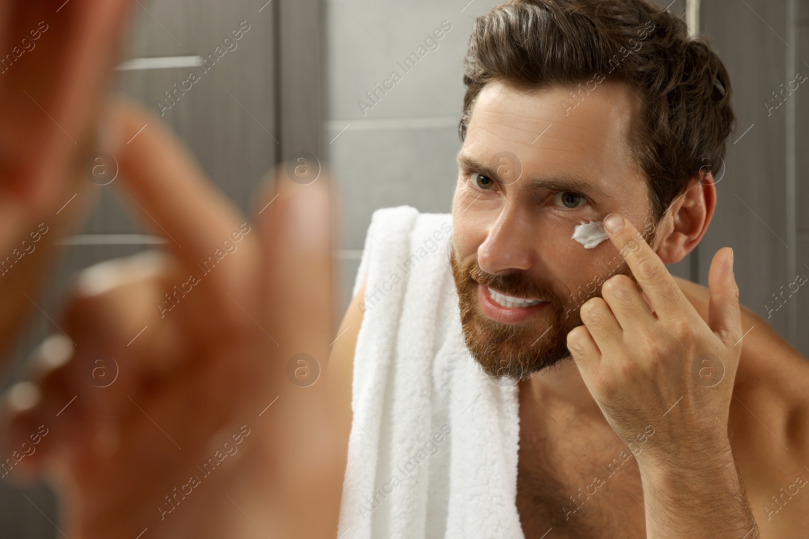 Photo of Handsome man applying cream on face in bathroom near mirror