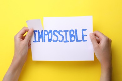 Photo of Motivation concept. Woman making word Possible from Impossible by tearing paper on yellow background, top view