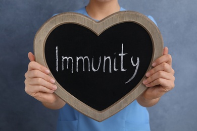 Woman holding heart shaped blackboard with word Immunity on grey background, closeup