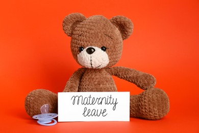 Photo of Toy bear, card with phrase Maternity Leave and pacifier on red background