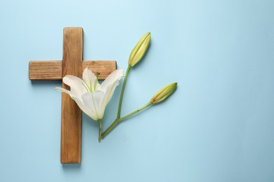 Photo of Wooden cross and lily flowers on light blue background, top view with space for text. Easter attributes