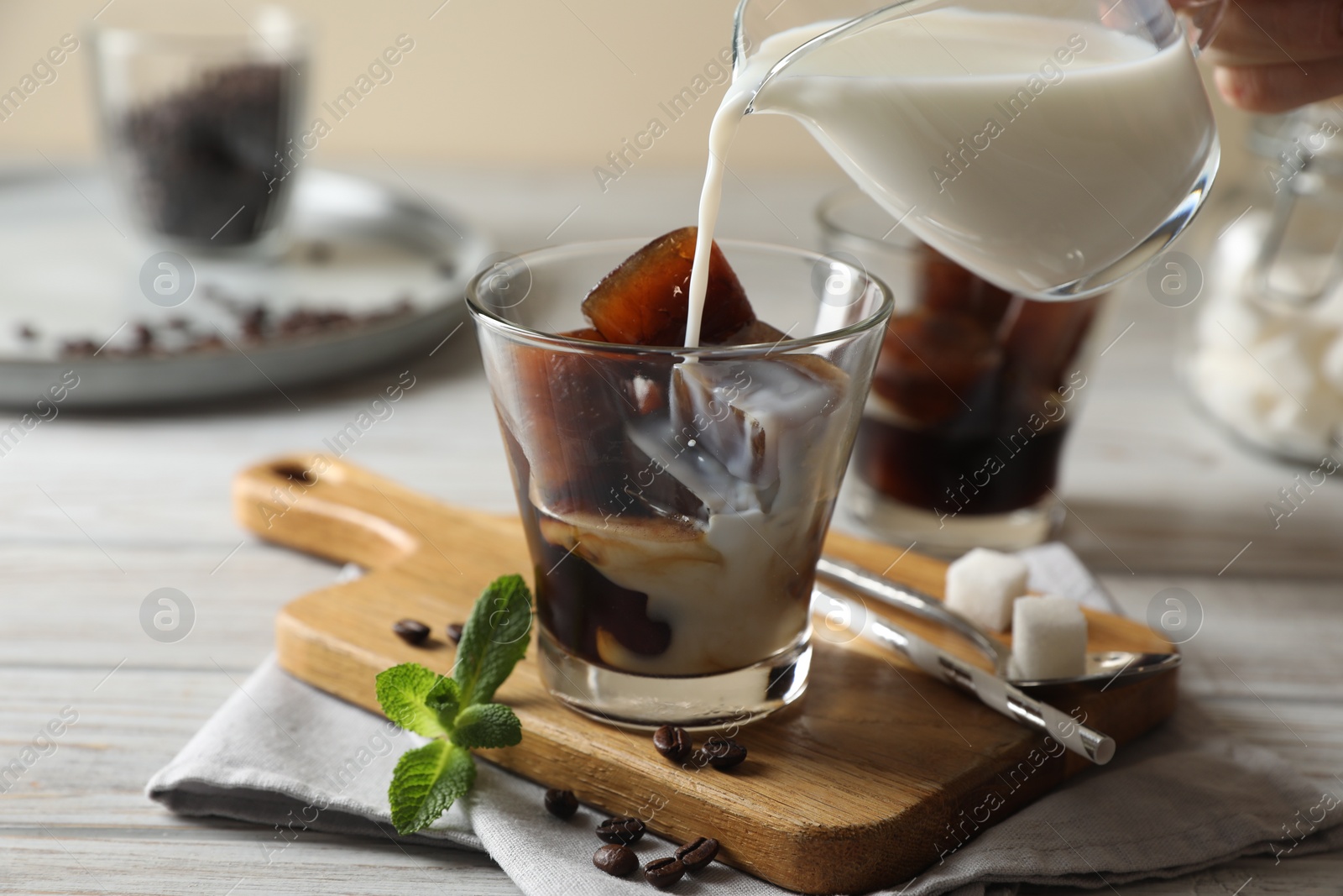 Photo of Pouring milk from jug into glass of delicious iced coffee, beans and mint on white wooden table