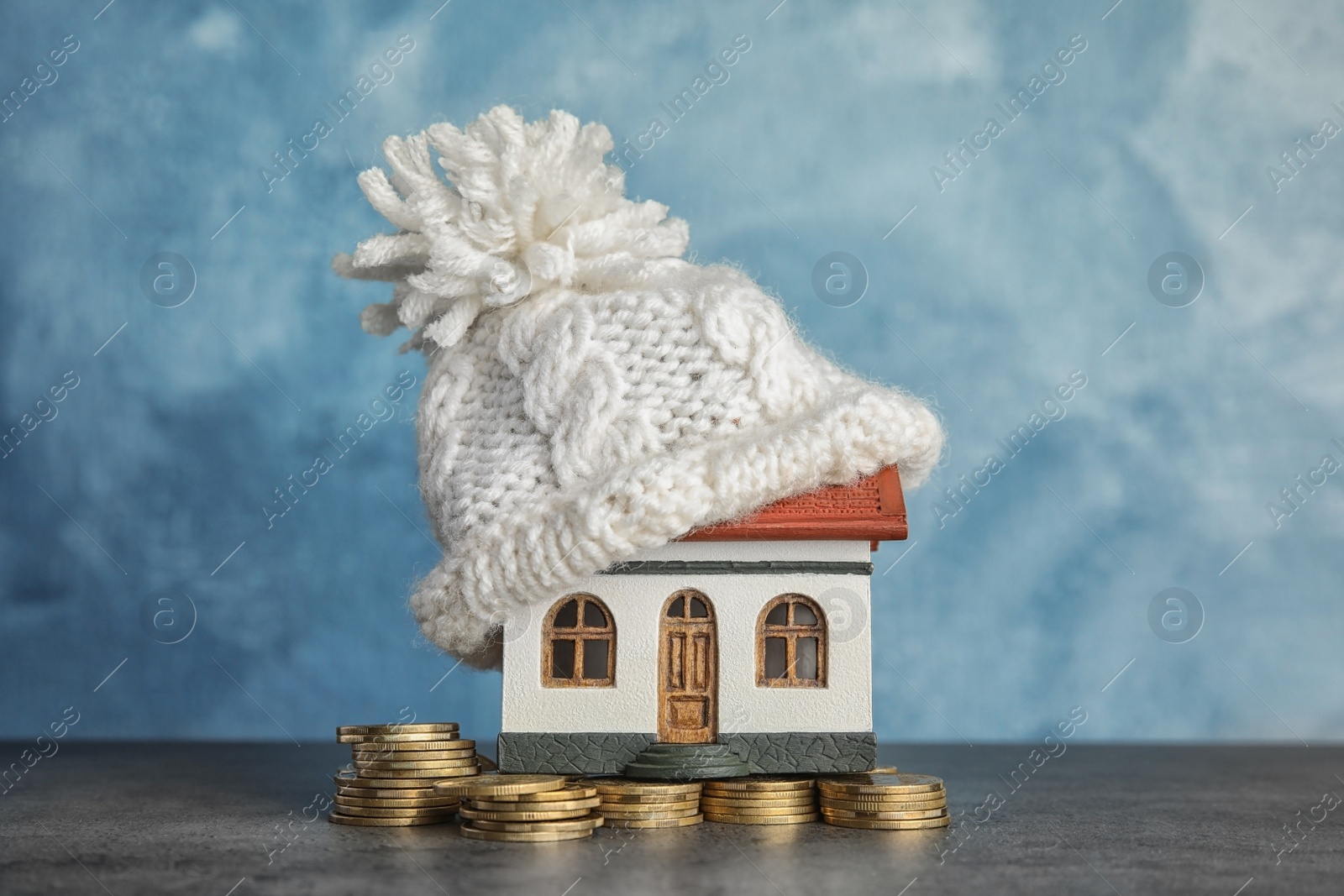 Photo of House model with knitted hat and coins on table against color background. Heating concept