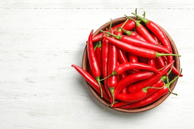 Photo of Wooden bowl with red hot chili peppers on white table, top view. Space for text