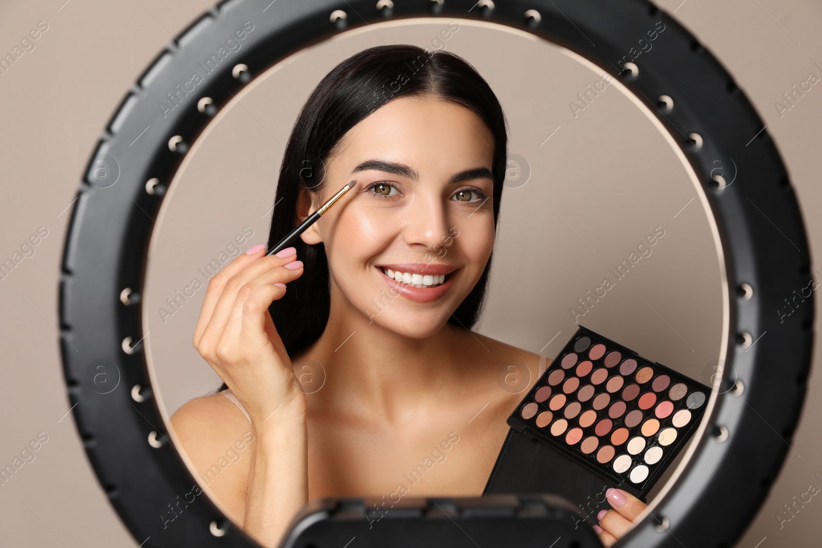 Photo of Beautiful young woman applying eye shadow with brush on beige background, view through ring lamp