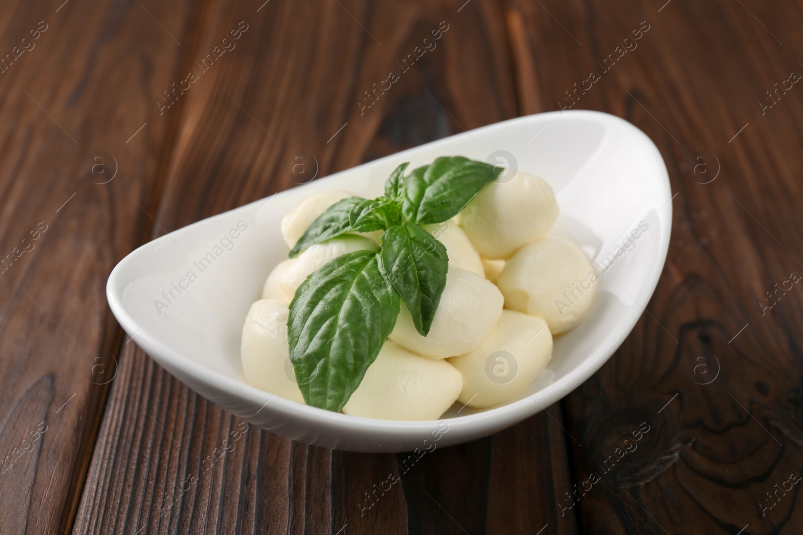 Photo of Tasty mozarella balls and basil leaves in bowl on wooden table, closeup