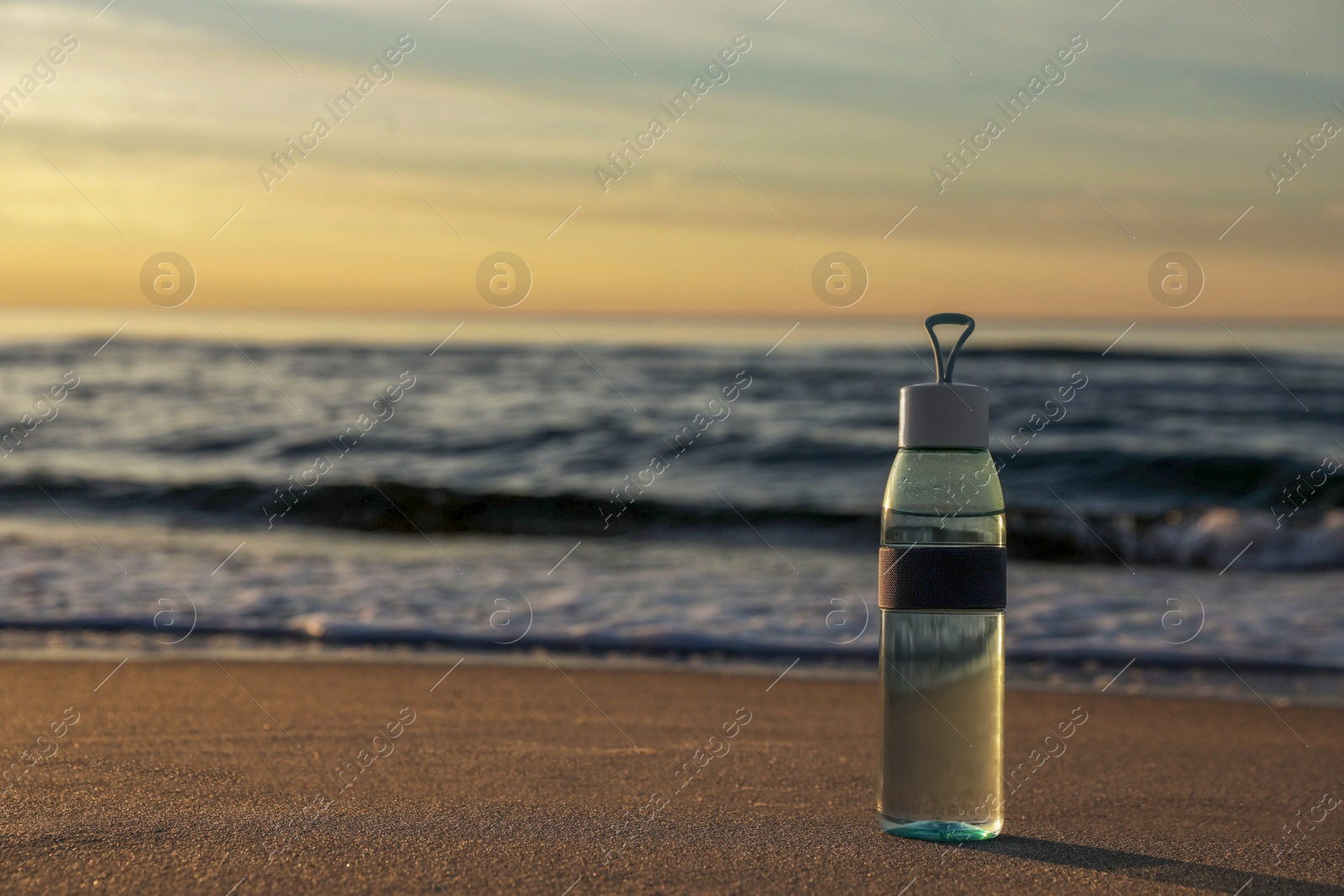 Photo of Glass bottle with water on wet sand near sea at sunset. Space for text