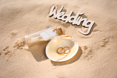 Photo of Shell with gold rings, invitation in glass bottle and word Wedding on sandy beach