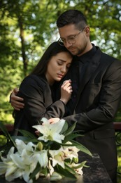 Photo of Sad couple mourning near granite tombstone with white lilies at cemetery. Funeral ceremony