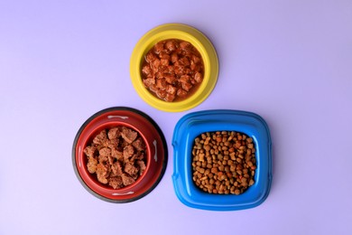 Wet and dry pet food in feeding bowls on violet background, flat lay