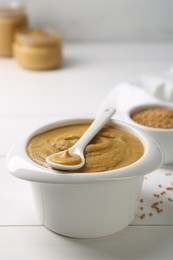 Photo of Spoon and bowl of tasty mustard sauce on white wooden table, closeup