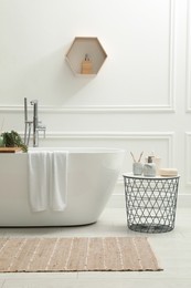 Modern ceramic bathtub and table with toiletries near white wall indoors