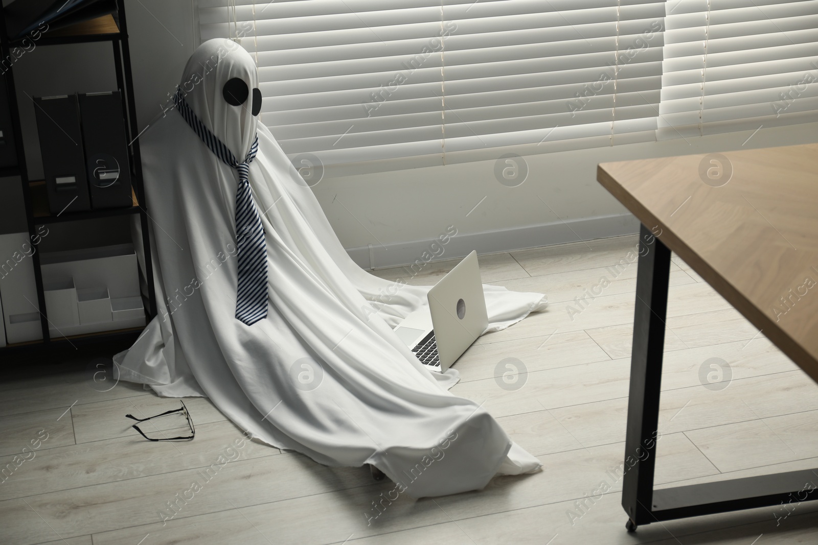 Photo of Overworked ghost. Man in white sheet with laptop on floor in office