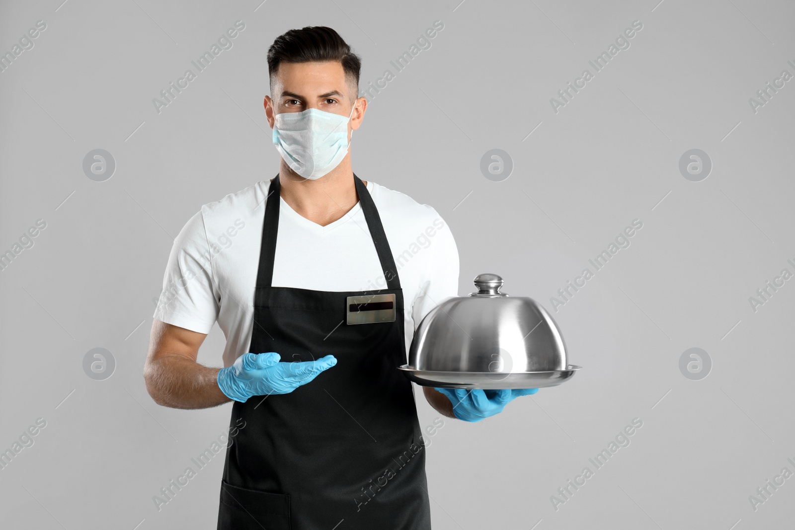 Photo of Waiter in medical face mask holding tray with lid on light grey background