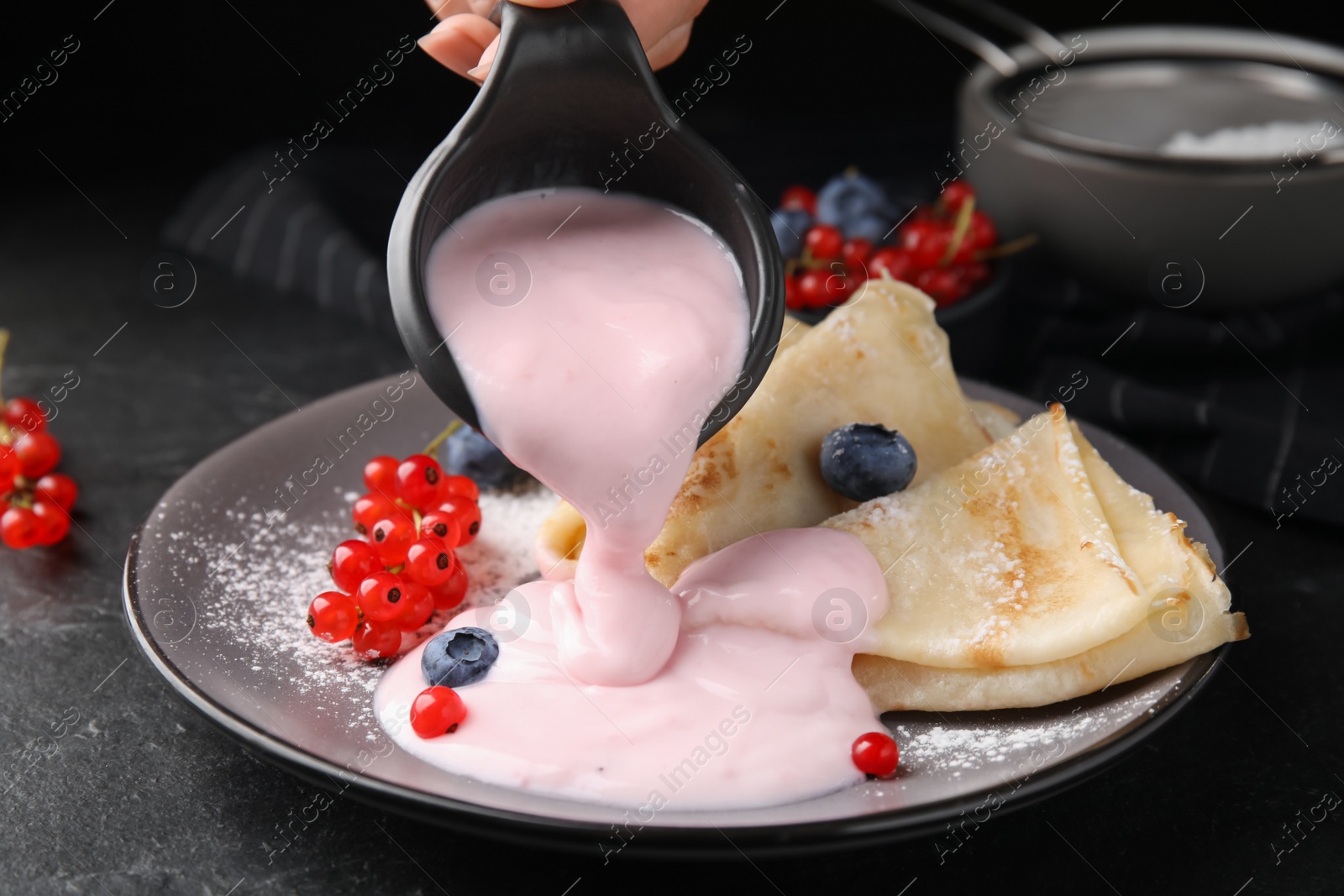 Photo of Woman pouring natural yogurt onto crepes with blueberries and red currants at table, closeup