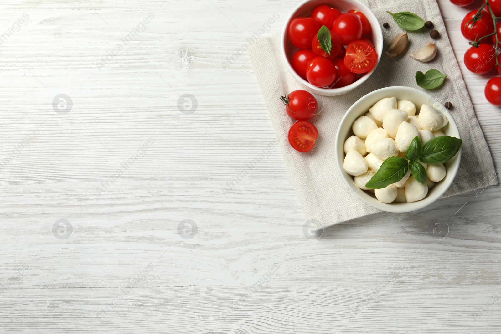 Photo of Delicious mozzarella balls in bowl, tomatoes and garlic on white wooden table, flat lay. Space for text