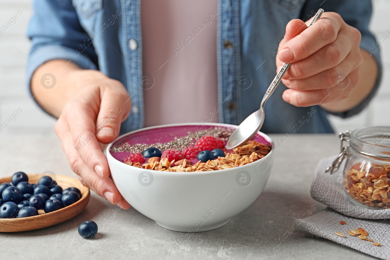 Photo of Woman eating acai smoothie with granola and berries at grey table, closeup