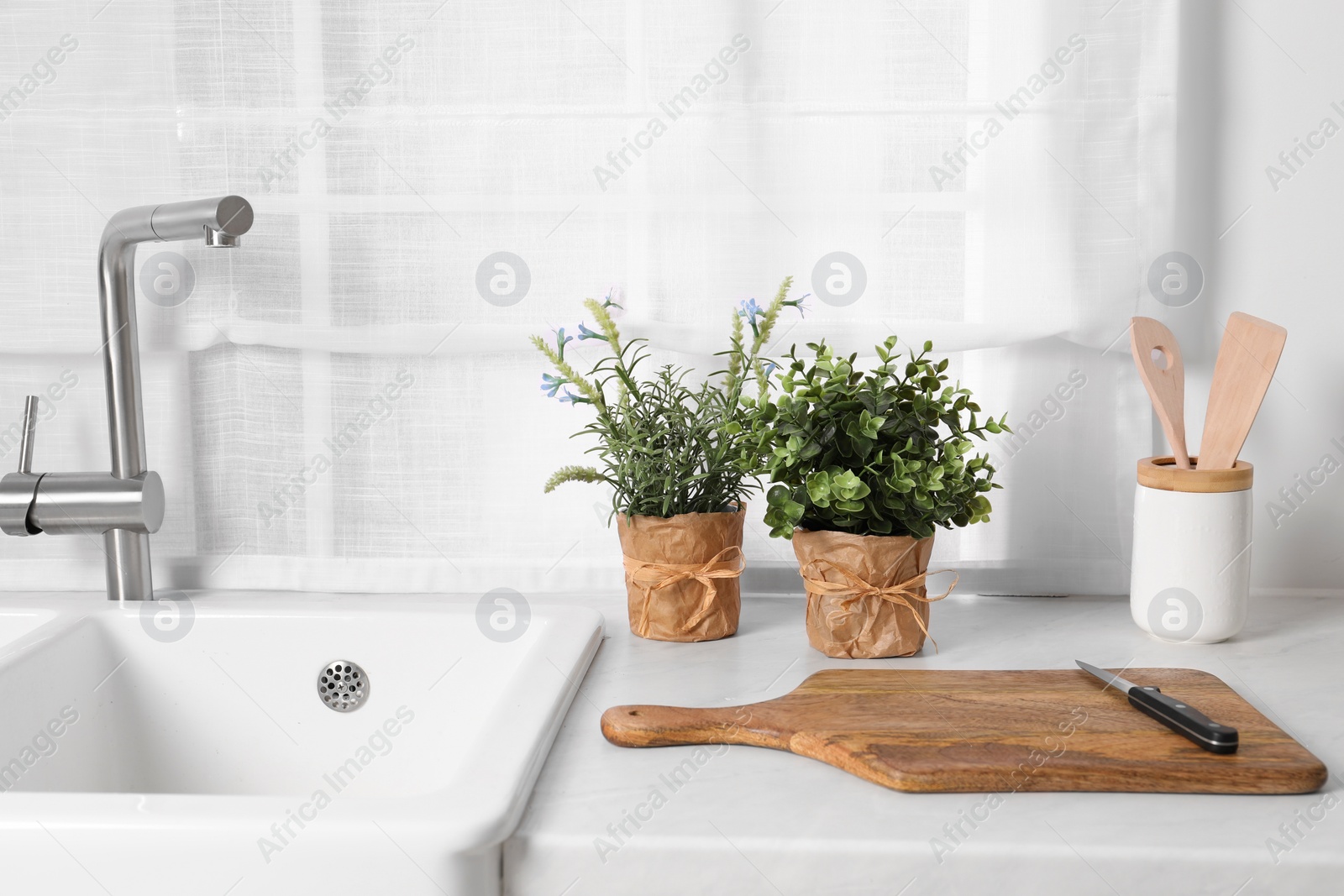 Photo of Artificial potted herbs, board and knife on white marble countertop in kitchen. Home decor