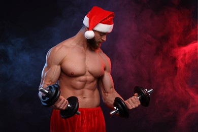 Muscular young man in Santa hat with dumbbells on color background, space for text