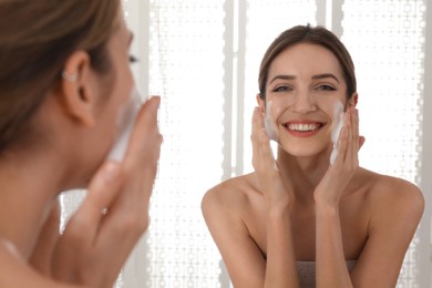 Young woman applying cleansing foam onto her face near mirror in bathroom