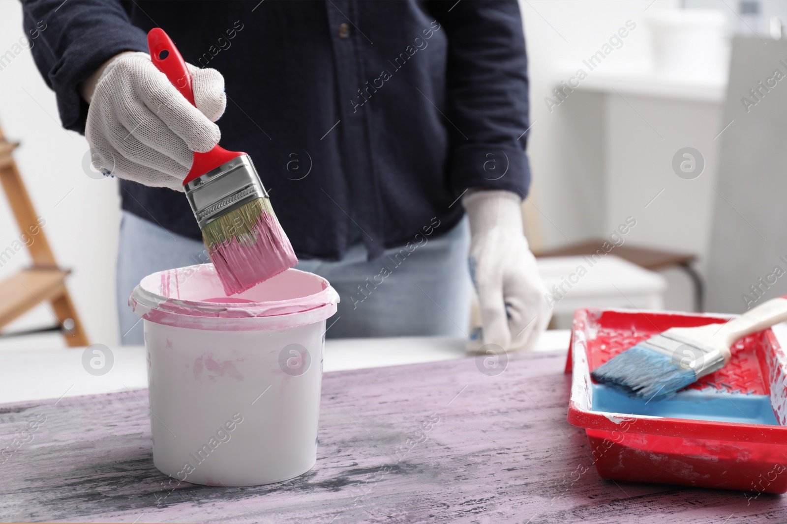 Photo of Woman dipping brush into bucket of pink paint indoors, closeup