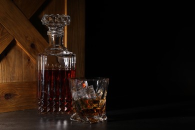 Photo of Whiskey in glass and bottle near wooden crate on dark table against black background. Space for text