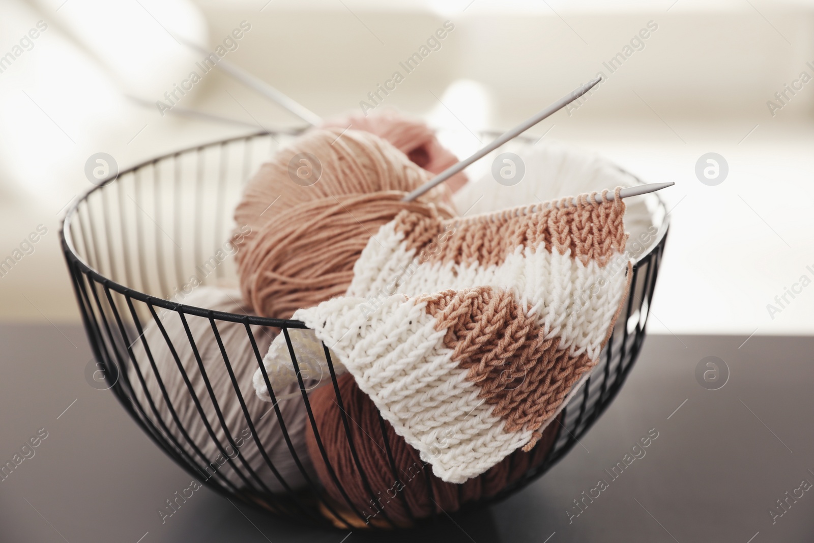 Photo of Yarn balls and knitting needles in metal basket on grey table against blurred background. Creative hobby