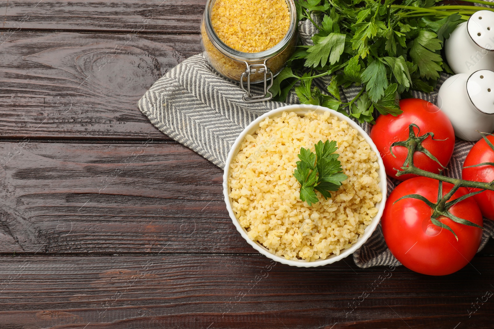 Photo of Delicious bulgur with parsley in bowl, tomatoes and spices on wooden table, top view. Space for text