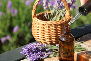 Photo of Dripping essential oil from pipette into bottle near lavender on wooden table outdoors, closeup. Space for text