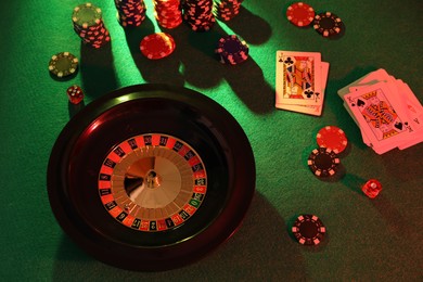 Photo of Roulette wheel with ball, playing cards and chips on green table, above view. Casino game