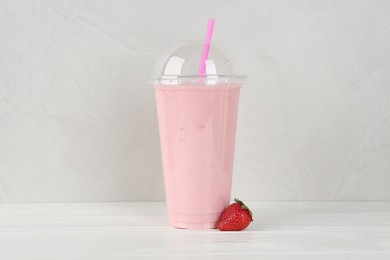 Plastic cup of tasty smoothie and fresh strawberry on white wooden table