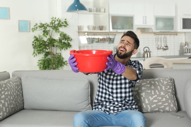 Young man holding plastic basin under water leakage from ceiling at home. Plumber service