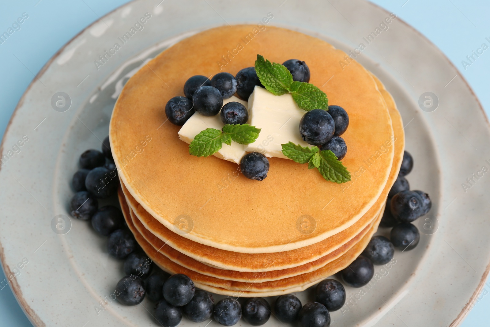 Photo of Stack of tasty pancakes with blueberries, butter and mint on light blue background, closeup