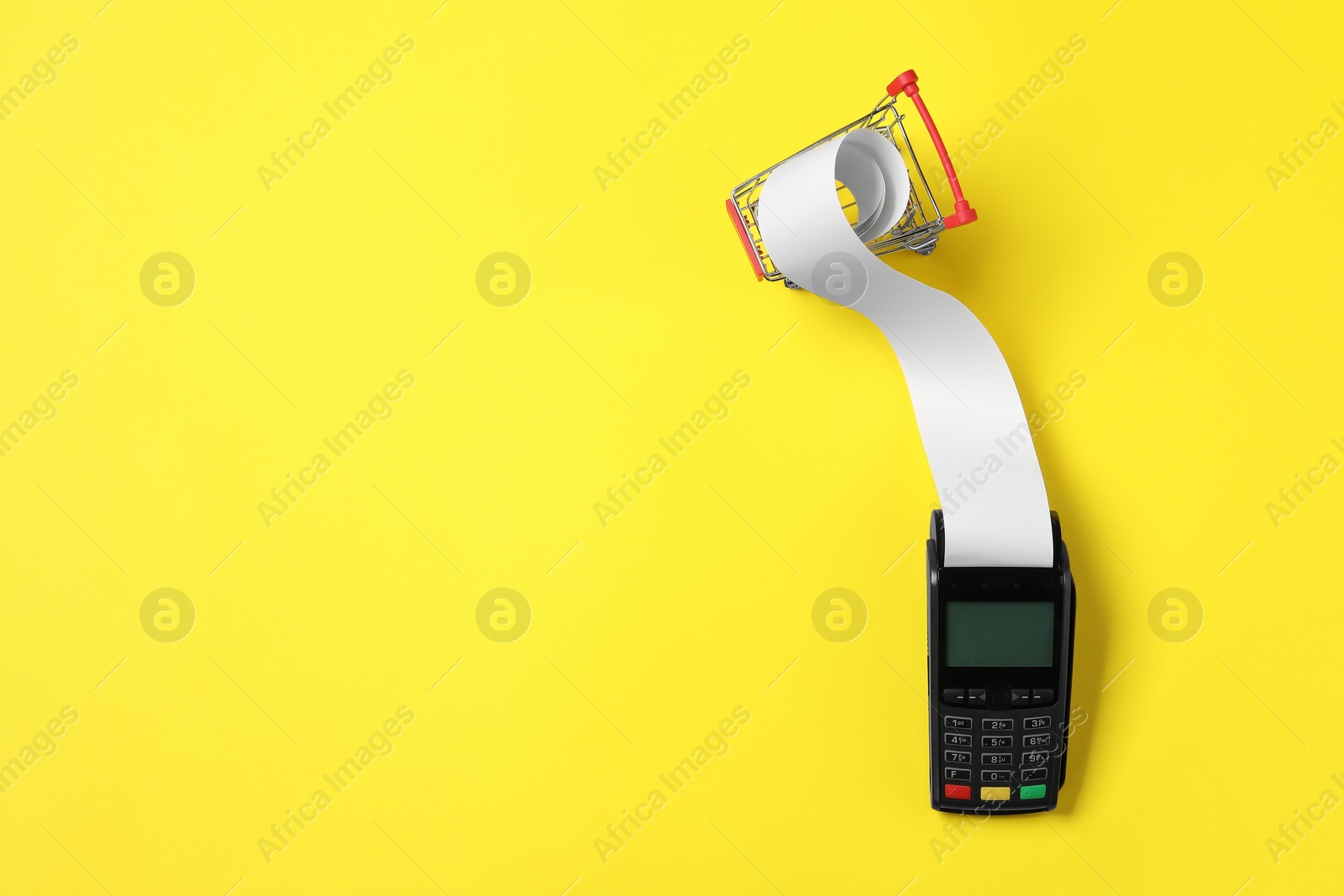 Photo of Payment terminal with thermal paper for receipt and small shopping cart on yellow background, flat lay. Space for text