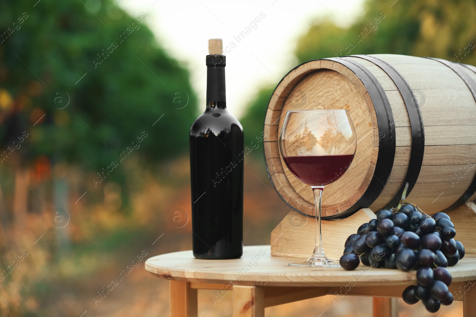 Photo of Bottle of wine, barrel and glass on wooden table in vineyard