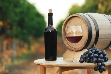Photo of Bottle of wine, barrel and glass on wooden table in vineyard