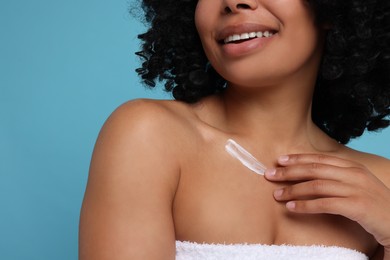 Young woman applying cream onto body on light blue background, closeup